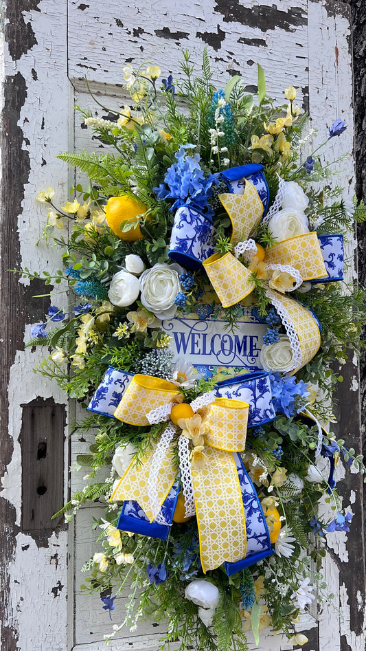 Full angled view of luxury lemon teardrop swag featuring layered blue Chinoiserie ribbon, yellow lattice ribbon, white open weave ribbon, ranunculus florals, lemon accents, mixed greenery and a decorative welcome sign in a dimensional handcrafted Gardenbug Designs arrangement.