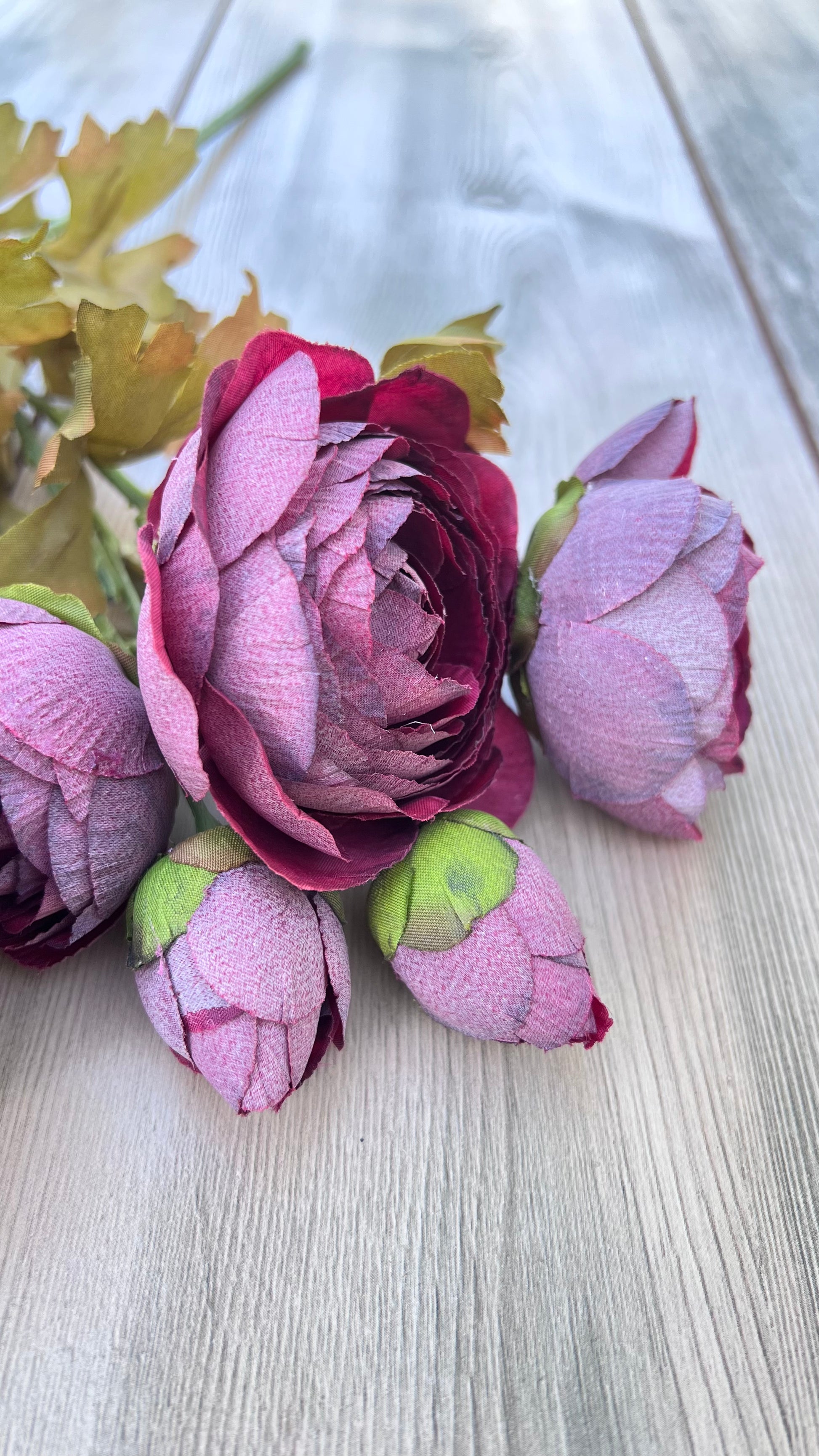 Our artificial Ranunculus Bundle in Burgundy featuring richly layered blooms, realistic buds, and soft foliage laying upon a wooden surface. Sourced by Gardenbug Designs.