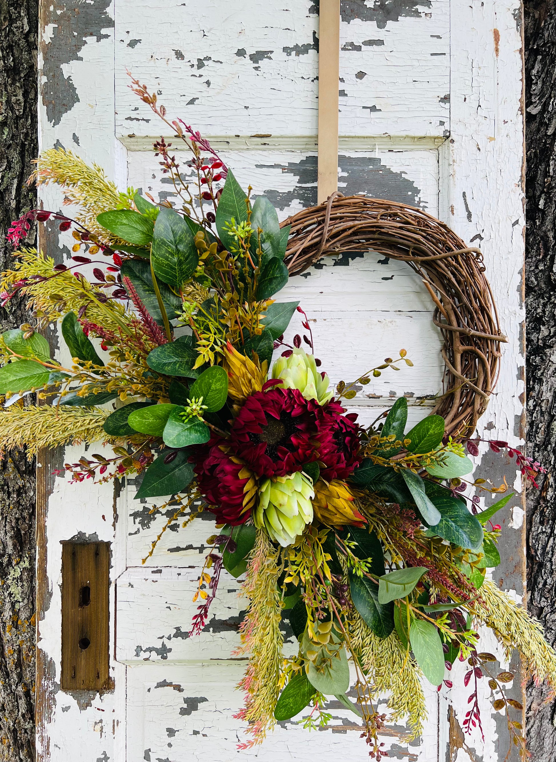 Autumn’s Garden botanical wreath design featuring burgundy sunflowers, green artichokes, and layered fall greenery styled on a rustic white door | Gardenbug Designs.