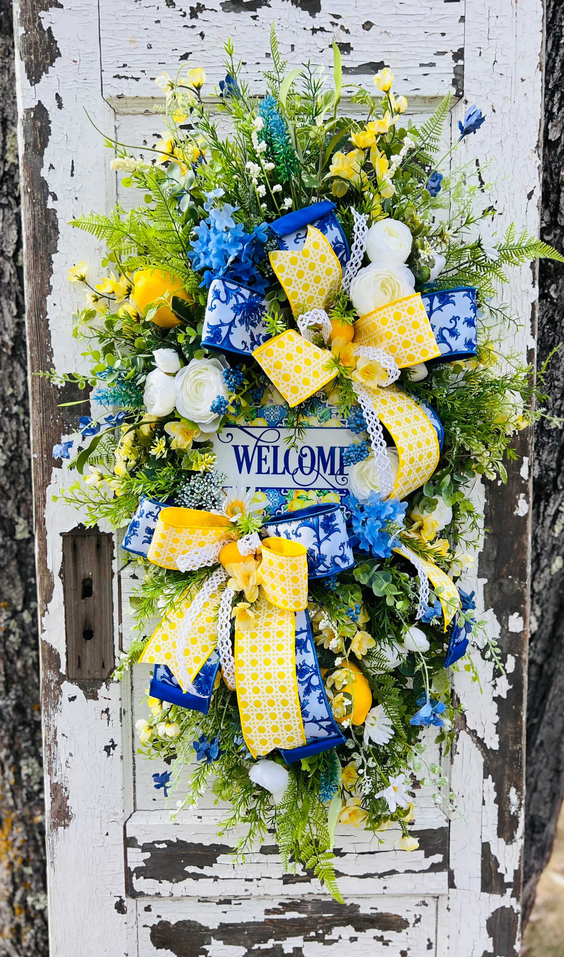 Luxury lemon citrus teardrop swag with blue and yellow florals, ranunculus, daisies, greenery, and a layered Chinoiserie bow on an evergreen swag base featuring a decorative welcome sign, spring summer front door swag designed by Gardenbug Designs.