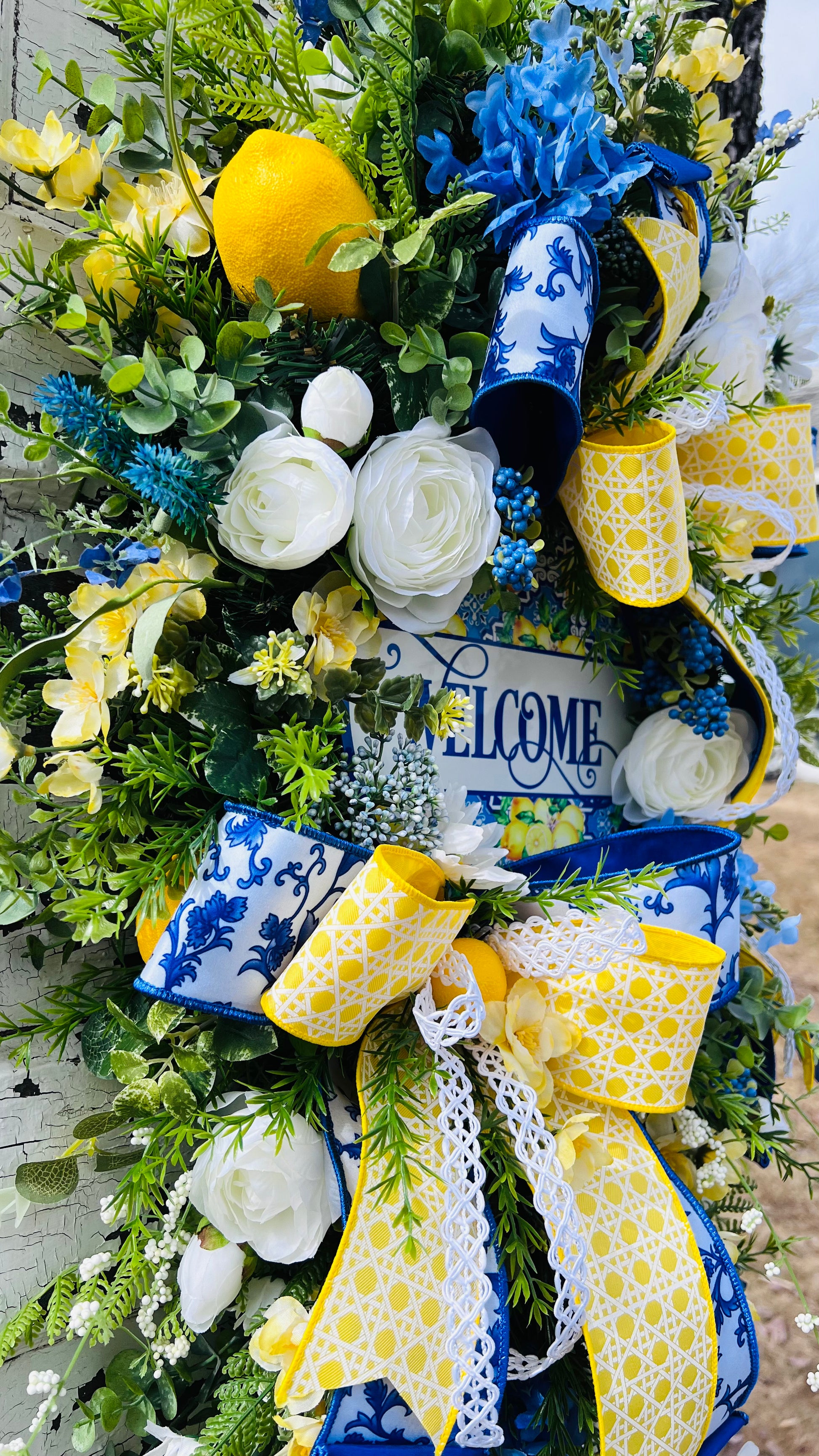 Close up side view of luxury lemon teardrop swag featuring layered designer ribbon, ranunculus florals, lemon accents and botanical greenery surrounding the welcome sign in a dimensional handcrafted Gardenbug Designs summer door arrangement.