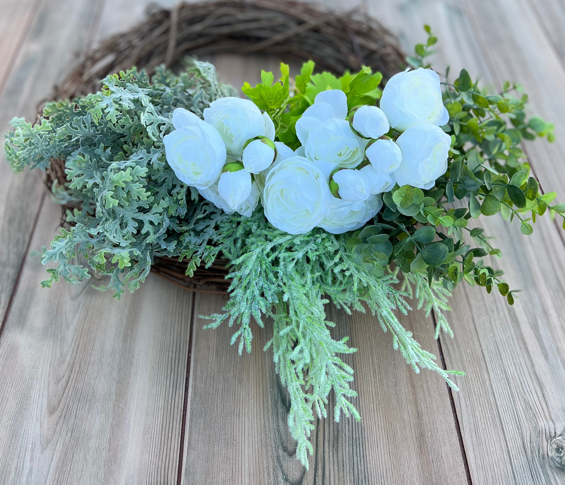 White ranunculus bundles placed on a grapevine wreath base with mixed greenery, showing bloom placement and layering